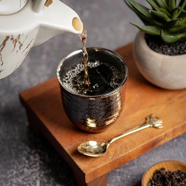 Hot tea being poured from a white teapot into a textured black and gold cup on a wooden tray with a gold spoon, loose tea leaves, and a small potted plant.