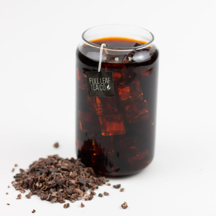 A glass jar filled with dark brewed Organic Cocoa Pu-Erh tea over ice, with a Full Leaf Tea Co. tag hanging from the rim, and a small pile of cocoa nibs scattered beside it on a white background.