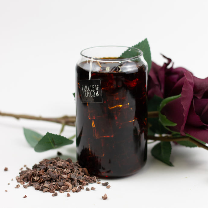 Glass of tea with a Fullleaf Cacao label, surrounded by cacao nibs and a rose on a white background.