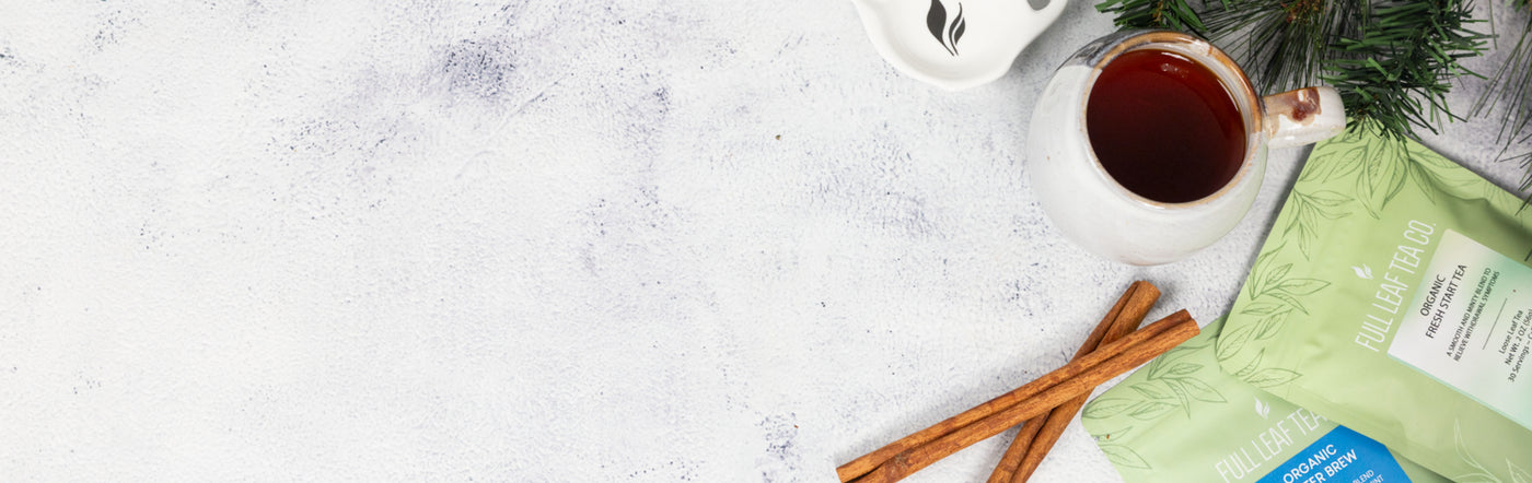 Some packages of tea next to a couple cinnamon sticks, a wooden spoon filled with the loose leaf blend, and some pine tree limbs, all on top of a marble surface.