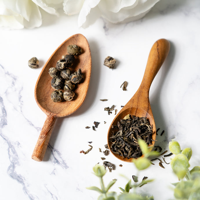 Two wooden spoons filled with black tea leaves on a marble surface with white flowers and green leaves.