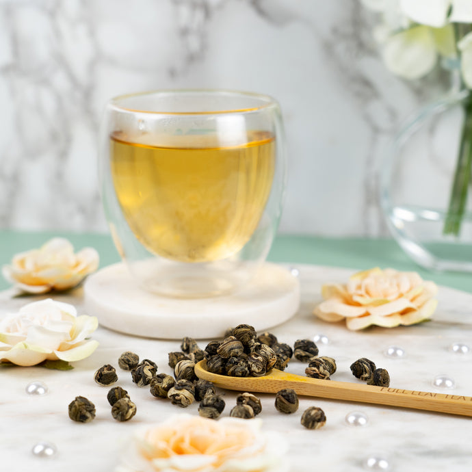 Jasmine pearls in front of a cup of Jasmine pearl tea with a white background and marble counter. 