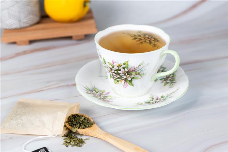 Cup of brewed tea in a floral china teacup and saucer, with a wooden spoon of loose leaf tea and a tea bag on a marble surface.