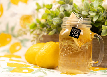 Mason jar with Lemon Myrtle tea and lemons on a floral tablecloth with leaves in the background. 
