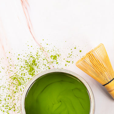 Ceramic bowl filled with vibrant green matcha tea beside a bamboo whisk on a white marble surface sprinkled with matcha powder.