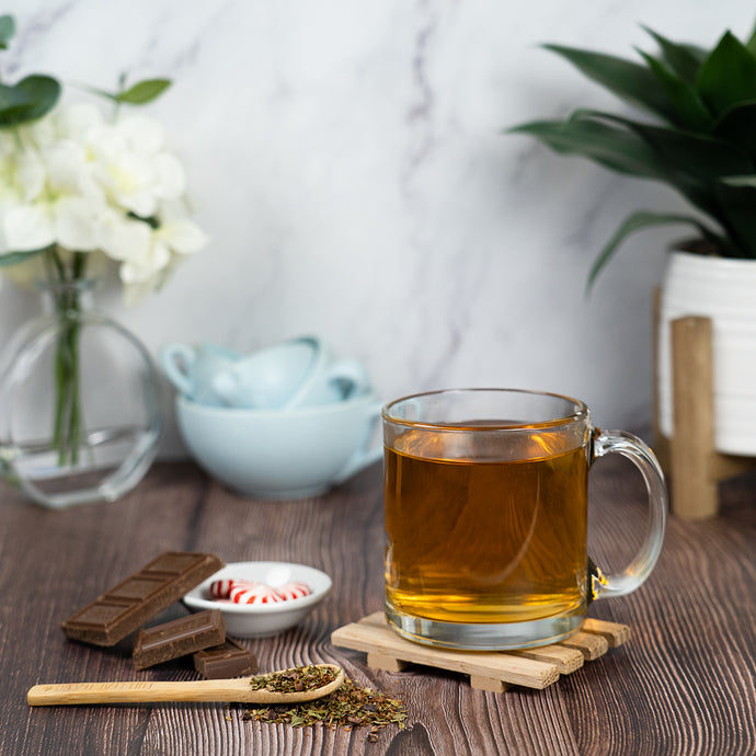 Glass mug of tea on a wooden coaster with chocolate and flowers in the background