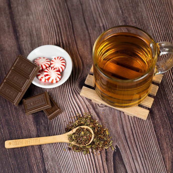 Glass of tea with a wooden spoon of herbs, chocolate bar, and peppermint candies on a wooden surface.