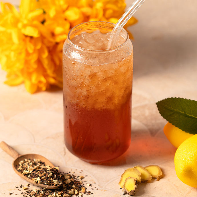 Glass of iced tea with a straw, surrounded by lemons, ginger, and a wooden spoon with tea leaves on a light background.