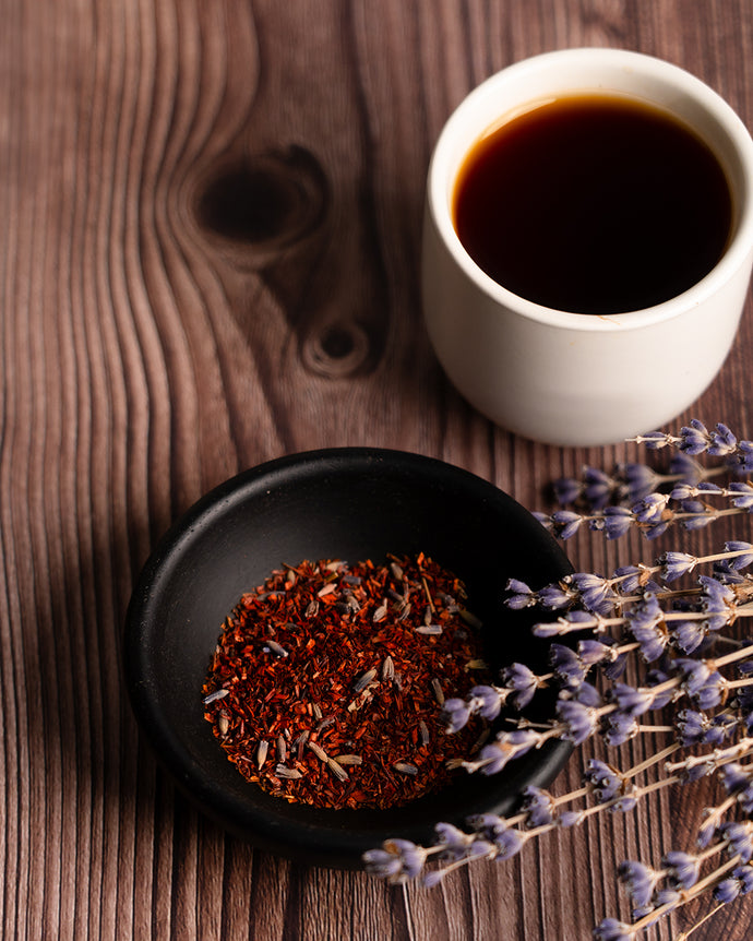 White cup of black coffee with a bowl of dried red rooibos tea and lavender on a wooden surface.