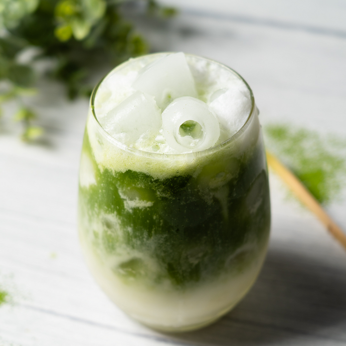 Iced matcha latte with creamy layers of green tea and milk, topped with ice cubes in a clear glass on a light wooden surface.