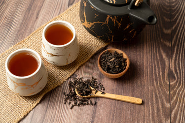 Two cups of brewed oolong tea beside a black and gold teapot on a wooden table with loose oolong tea leaves and a bamboo spoon engraved with 'Full Leaf Tea Co.