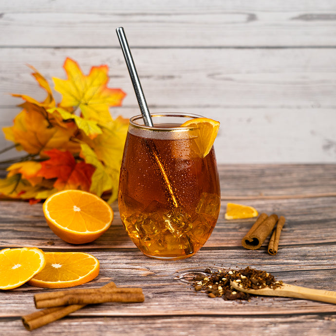 Glass of iced tea with a straw, surrounded by orange slices and cinnamon sticks on a wooden surface.