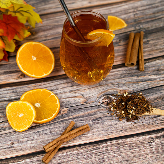 Glass of iced orange cinnamon spice tea with oranges, cinnamon sticks, and a spoonful of tea leaves on a wooden surface.