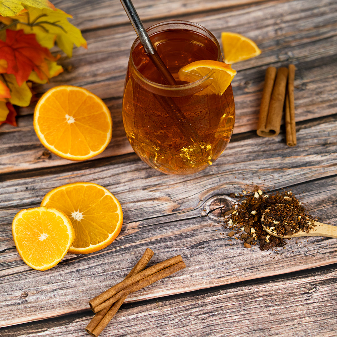 Glass of iced tea with oranges, cinnamon sticks, and a spoonful of tea leaves on a wooden surface.