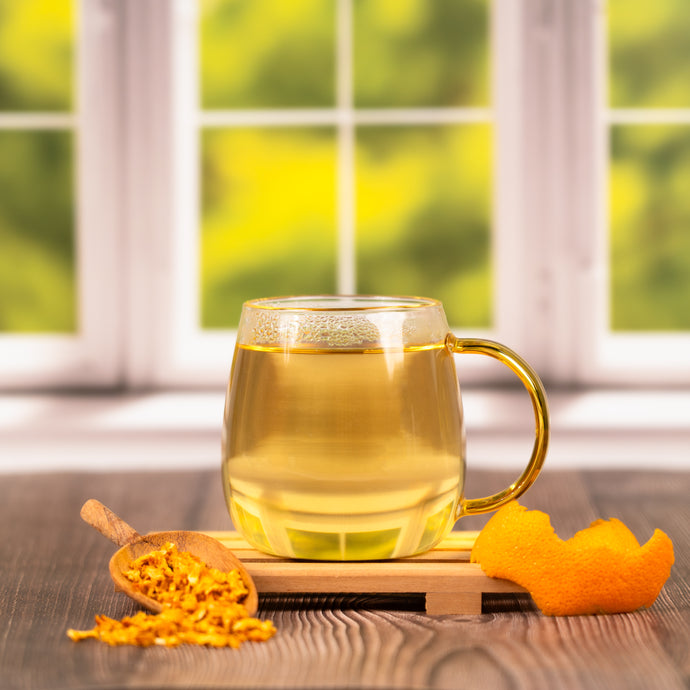 Glass mug with orange peel tea on a wooden coaster, surrounded by orange peels and a spoonful of dried fruit, with a blurred green outdoor background.