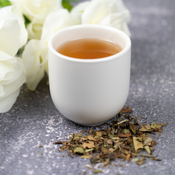 White cup of Pai Mu Tan tea with dry leaves and white flowers on a gray surface