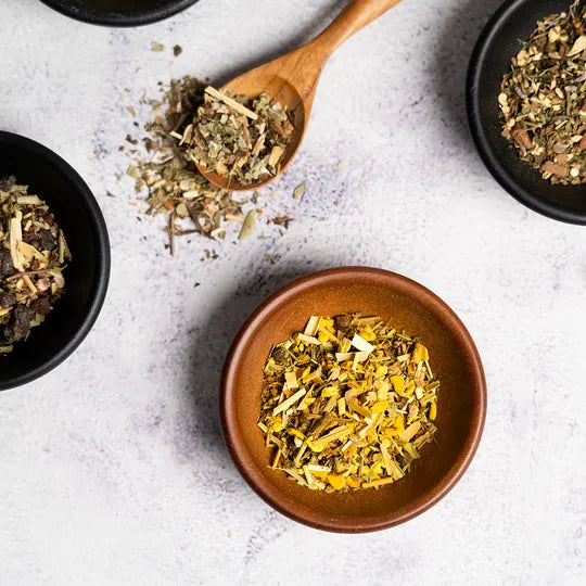 Assorted dried herbs displayed in small bowls and a wooden spoon on a light surface, showcasing the ingredients used in Organic Pain Relief Tea.