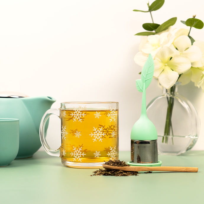 Glass mug of Organic Bloat Relief Tea with loose leaves, a green tea infuser, and a teapot beside white flowers.