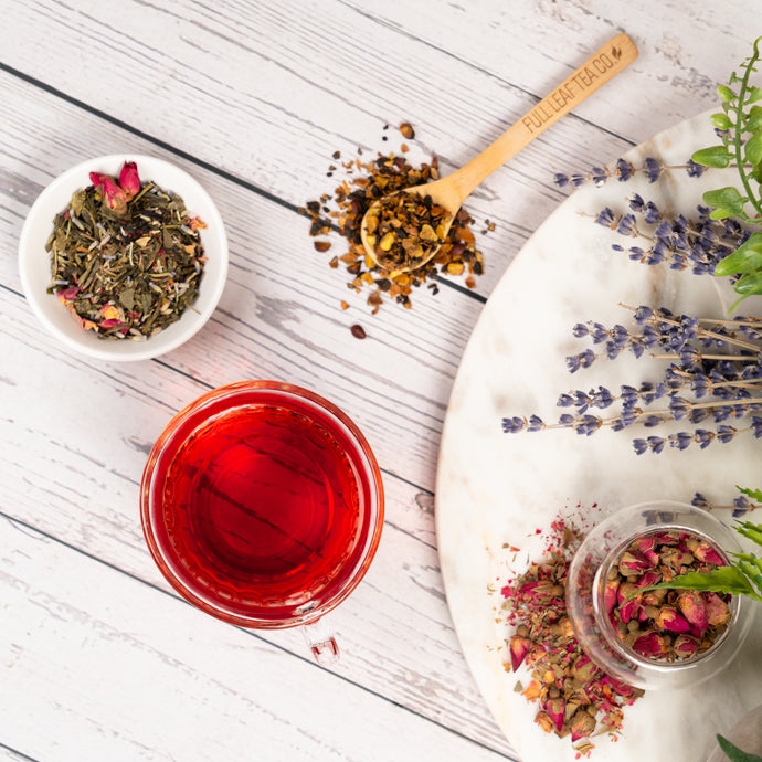 Tea-making setup with a cup of pink tea, dried herbs, and a wooden spoon on a white wooden surface.