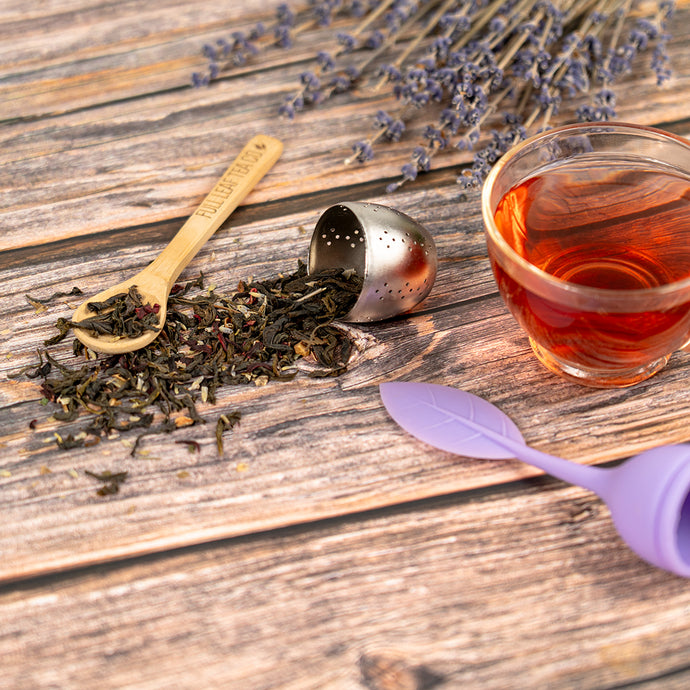 Tea leaves, a tea infuser, and a glass of tea on a wooden surface with lavender flowers.