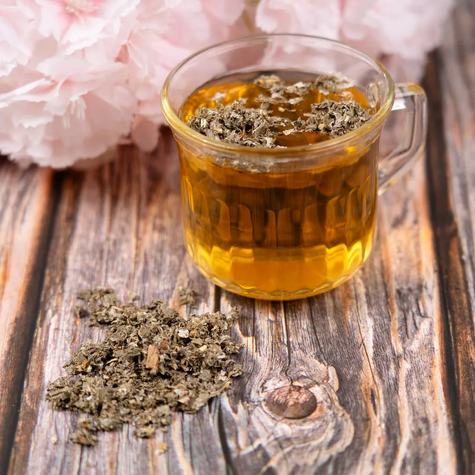 Glass mug of tea with dried raspberry leaf on a wooden surface, pink flowers in the background