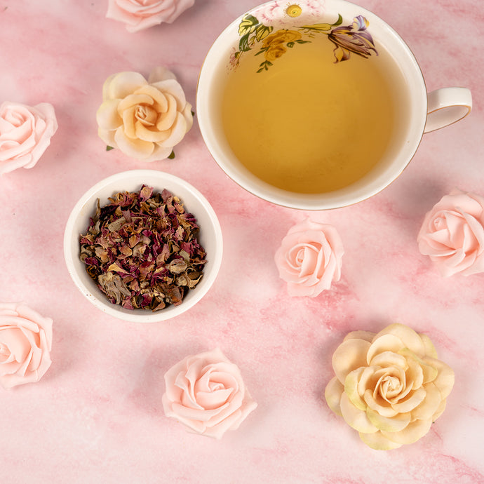 A cup of Rose Petal Tea in a white mug with a bowl of rose petals beside it and the surface is marble with rose petals on it. 