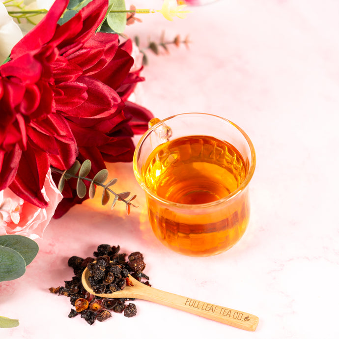 Glass of rosehips tea with a spoonful of rosehip leaves and red flowers on a light pink background