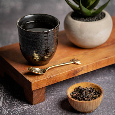 A black ceramic cup of Organic Royal Blend tea accompanied by a metal spoon, and a potted succulent on a wooden table, next to a small wooden bowl of the loose leaf tea blend.
