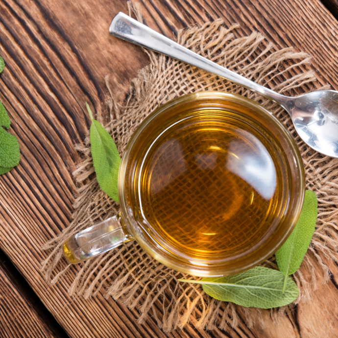 Clear glass mug filled with a sage tea, on a rustic wooden surface with a spoon and green leaves.