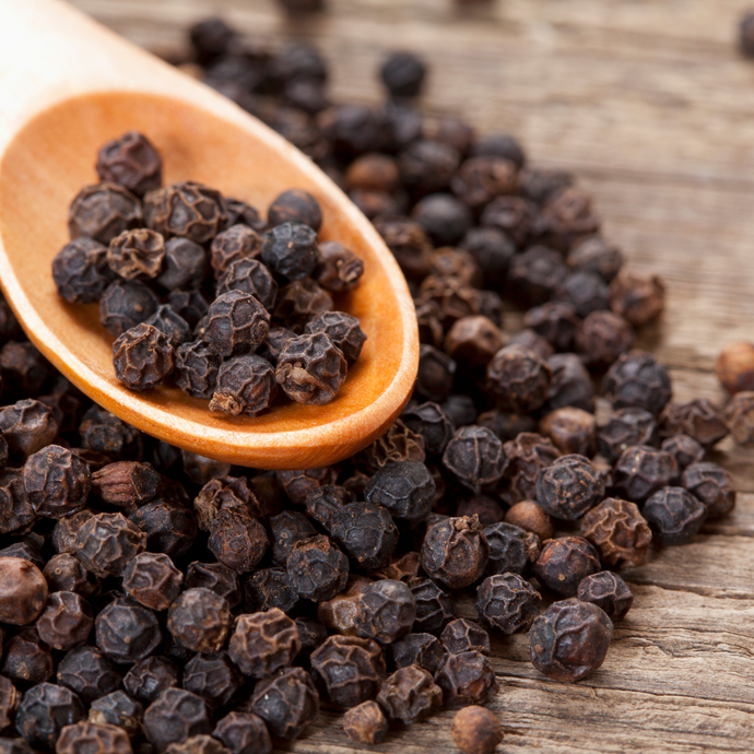 Close-up of whole black peppercorns with a wooden spoon on a rustic wooden surface.