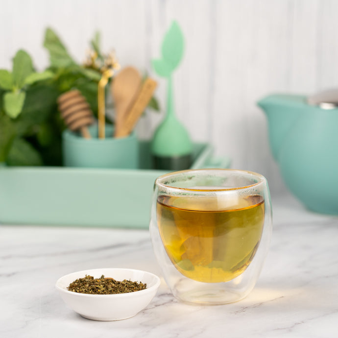Glass of spearmint tea with a small bowl of spearmint leaves on a marble surface, with a blurred background of kitchen utensils and plants.