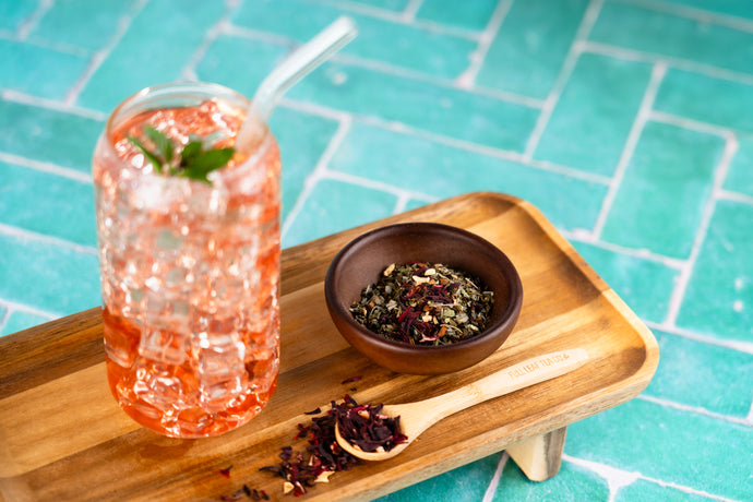 Glass of pink drink with ice and mint, bowl of tea leaves on a wooden tray against a blue tiled background