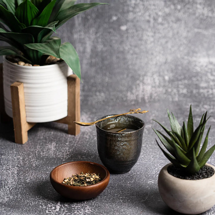 Tea-making setup with a cup, bowl of tea leaves, and potted plants on a gray surface.