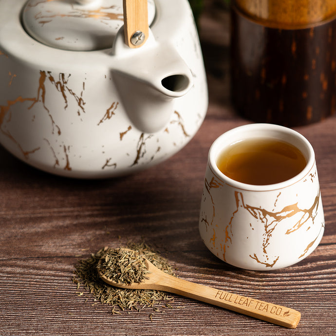 Tea set with marbled teapot, cup, wooden spoon, and thyme leaves on a wooden surface.
