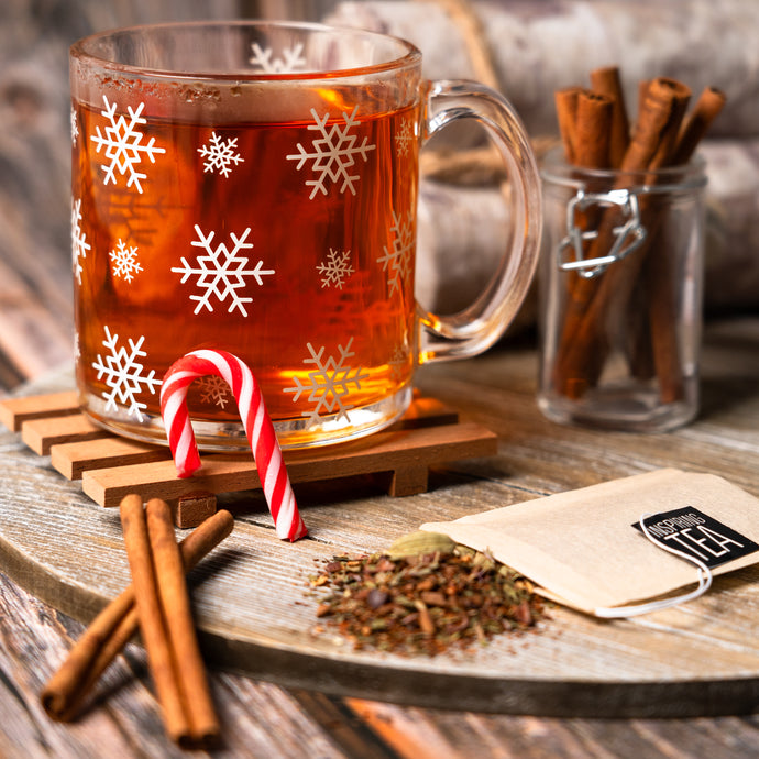 Glass mug with snowflake design filled with a winter brew tea, surrounded by cinnamon sticks and a candy cane on a wooden surface.