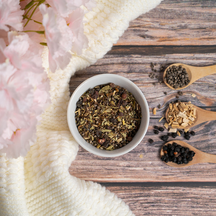 Tea leaves in a white bowl on a wooden surface with pink flowers and white fabric.