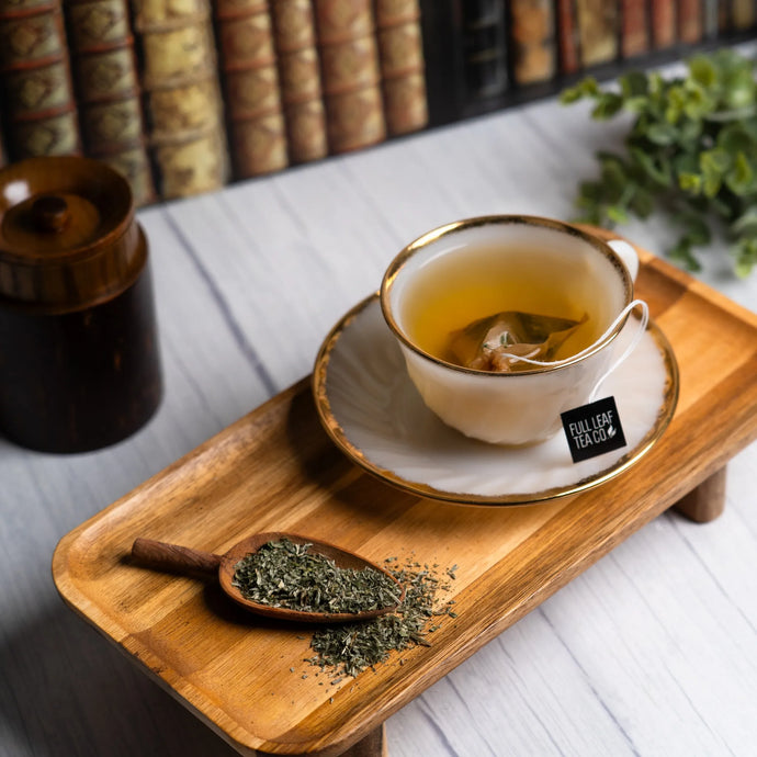 Tea cup with saucer on a wooden tray, surrounded by tea leaves and a teapot, with a blurred bookshelf in the background.