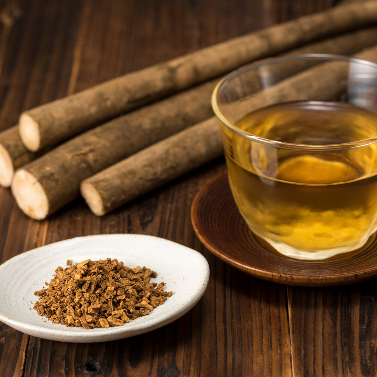 A clear mug of burdock root tea with burdock root in the background and burdock root ground up in a white saucer on a wooden surface. 