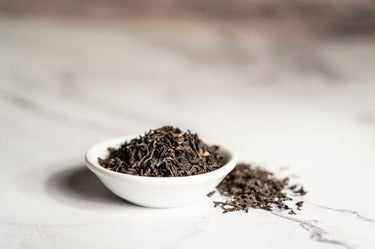 A small white bowl filled with loose-leaf black tea on a marble surface, with some tea leaves scattered beside it.