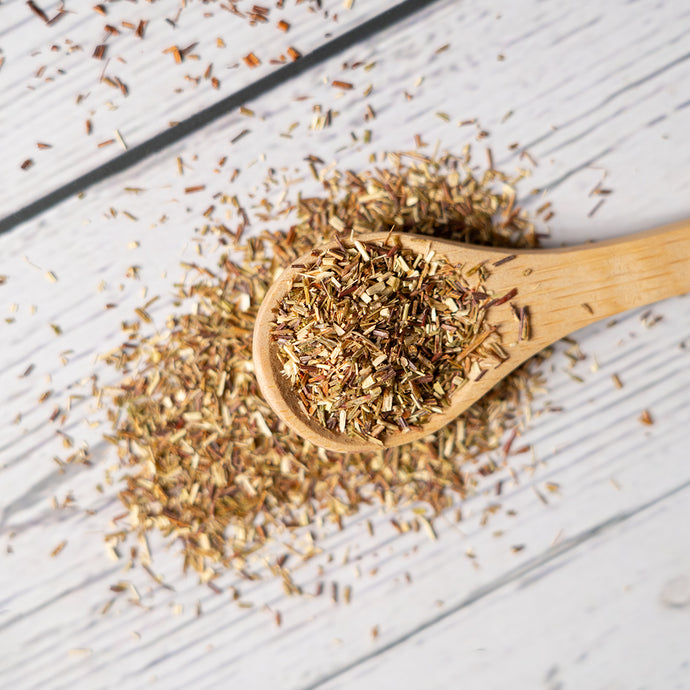 A wooden spoon filled with loose green rooibos tea leaves on a white wooden surface, with additional tea scattered around.