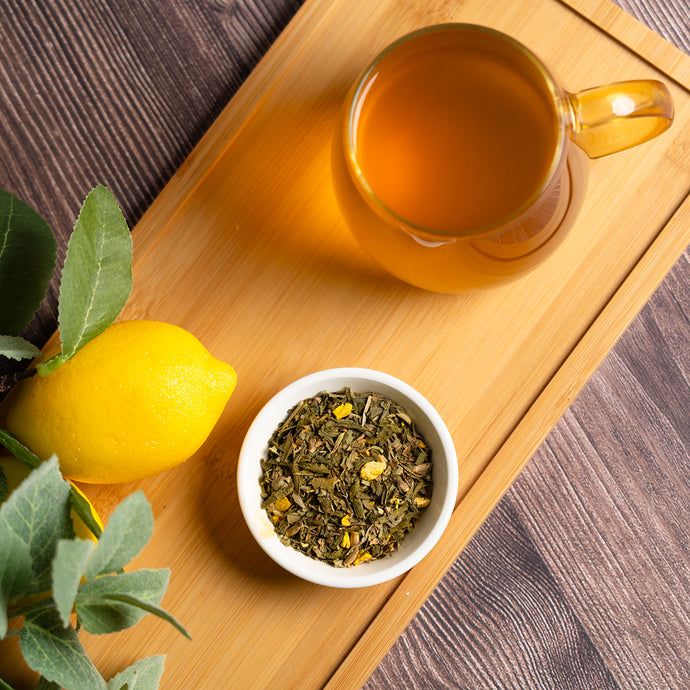 A glass mug of Organic Recovery Tea on a bamboo tray with a fresh lemon, green tea leaves mixed with lemon peel in a small white bowl, and leafy sprigs for garnish.