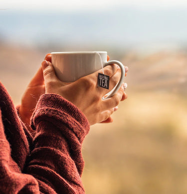 Person wearing a cozy maroon sweater holding a white mug of tea with a tag reading ‘Inspiring Tea,’ overlooking a soft blurred outdoor background.