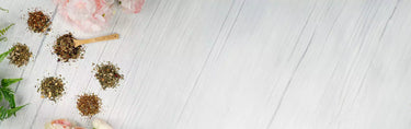 Assorted loose-leaf tea blends arranged on a white wooden surface with flowers and a wooden scoop.