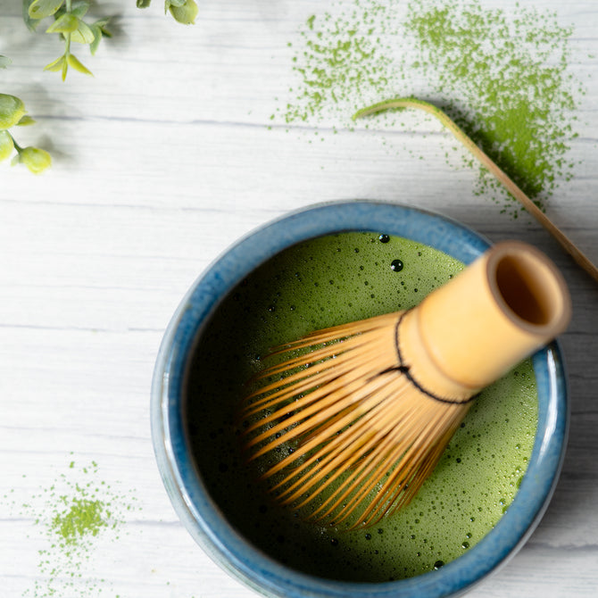 Bamboo matcha whisk resting in a blue ceramic bowl of freshly prepared green matcha tea, with matcha powder scattered on a white surface.