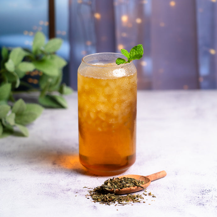 Glass of mint bliss tea with a mint leaf on a textured surface with dried tea leaves and greenery.