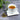 Cup of peppermint tea on a white saucer with a spoonful of dried peppermint leaves beside it, set on a gray textured surface with green foliage in the background.