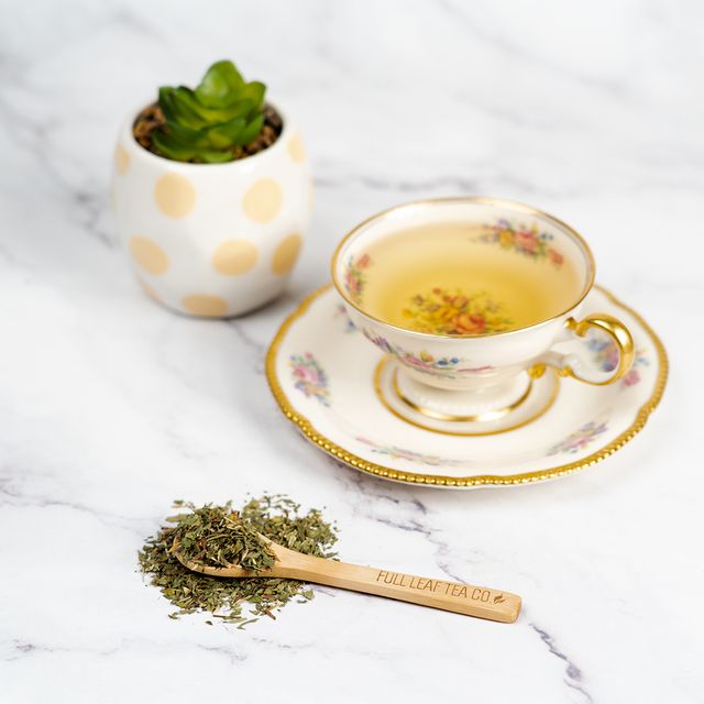Cup of Organic Red Clover tea in a floral porcelain teacup with loose tea leaves and a wooden spoon labeled Full Leaf Tea Co. on a white marble surface.