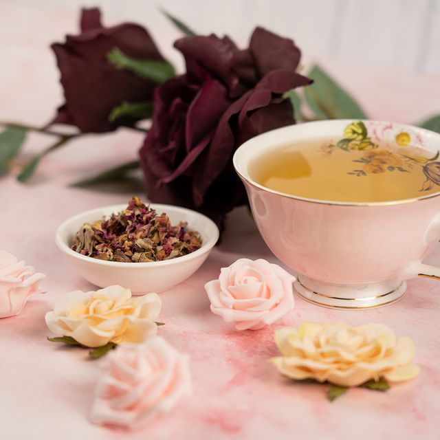 Cup of floral herbal tea beside dried rose petals and decorative roses on a pink surface.