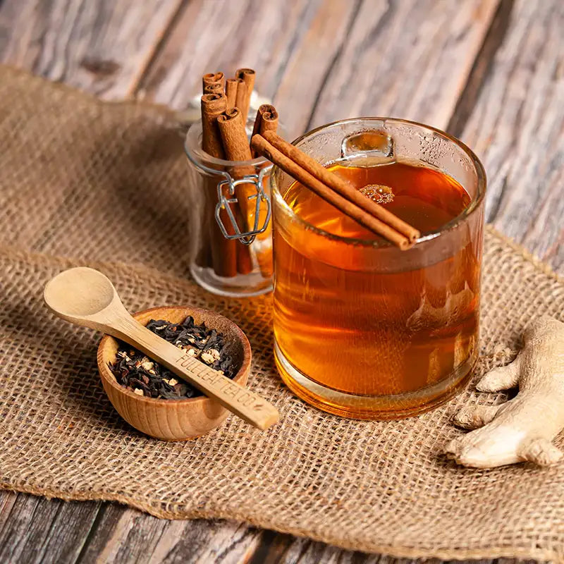 A glass of warm, aromatic tea with cinnamon sticks, fresh ginger root, and loose tea leaves on a wooden table with a rustic burlap cloth.