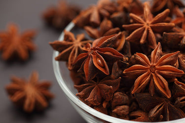 A clear glass bowl filled with star anise pods on a dark surface, with additional star anise scattered in the background.
